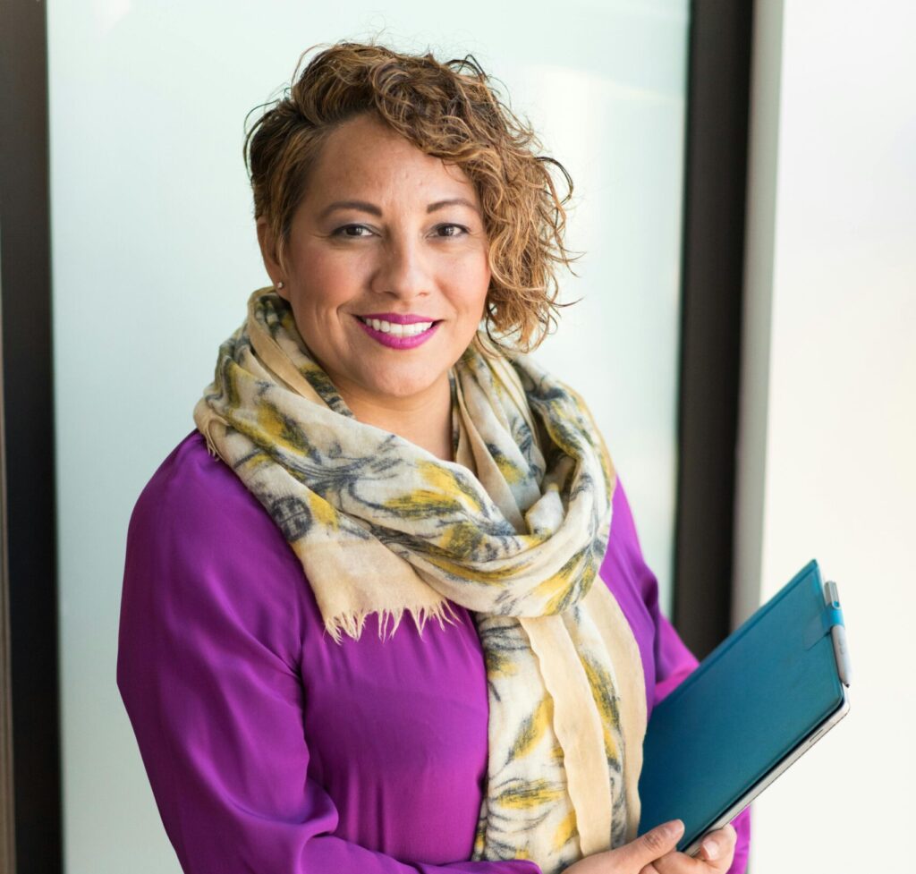 Smiling woman in a purple dress and scarf holding a tablet indoors.