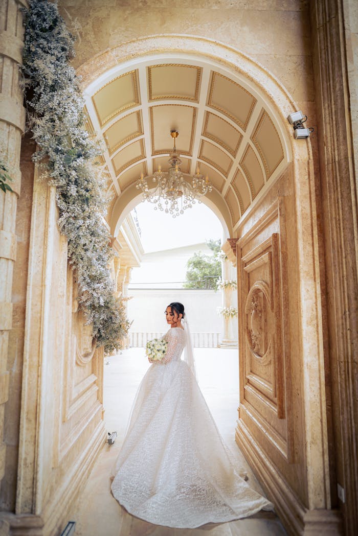 Bride in luxurious wedding dress standing in ornate archway, holding bouquet.